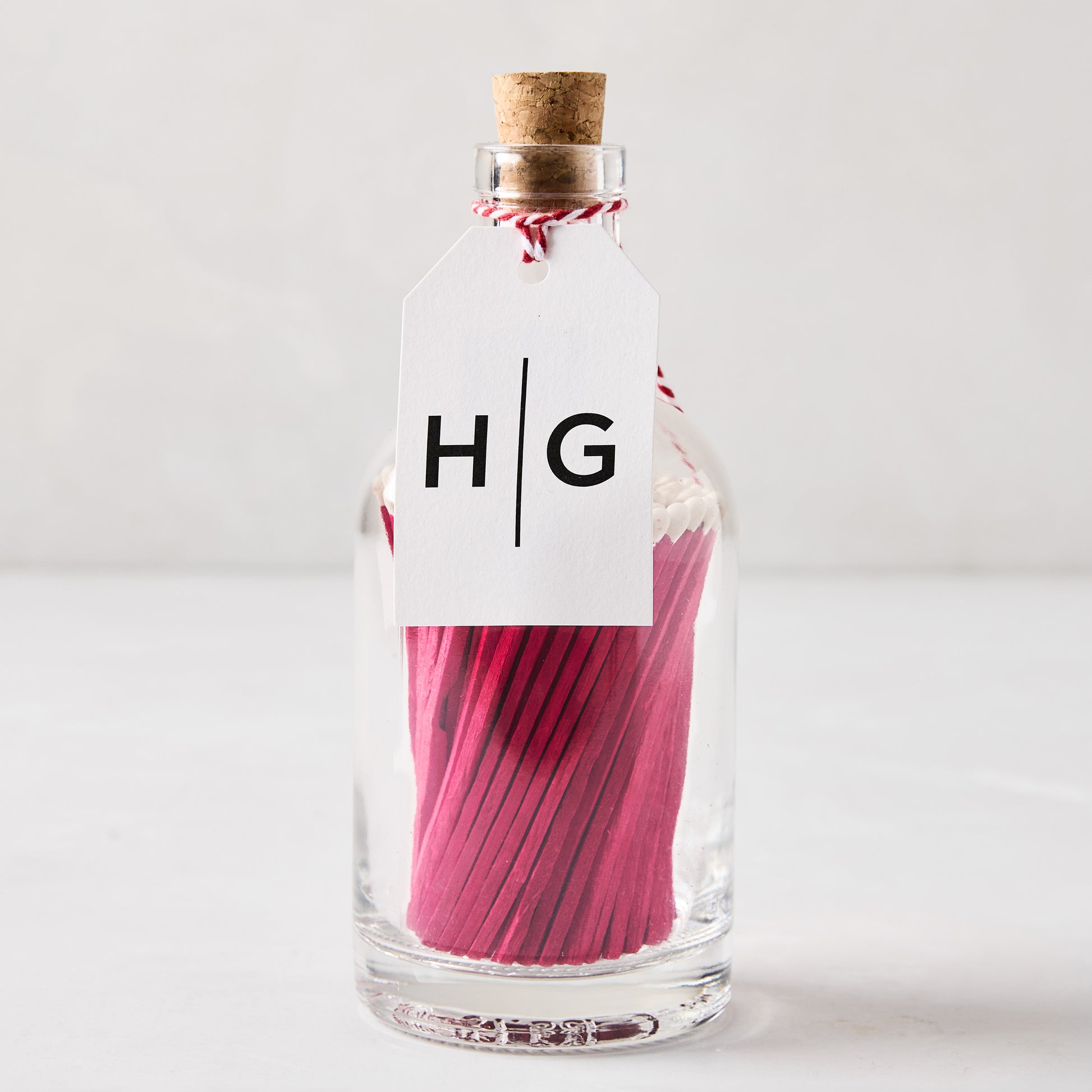 Glass bottle with cork stopper, filled with pink matches, and a white HG tag.