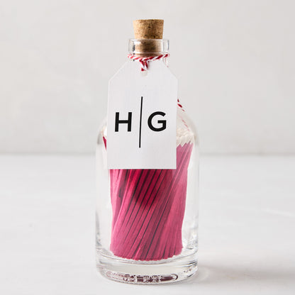 Glass bottle with cork stopper, filled with pink matches, and a white HG tag.