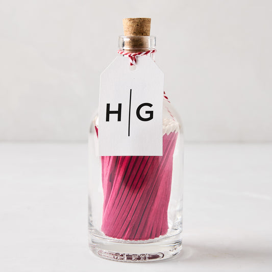 Glass bottle with cork stopper, filled with pink matches, and a white HG tag.