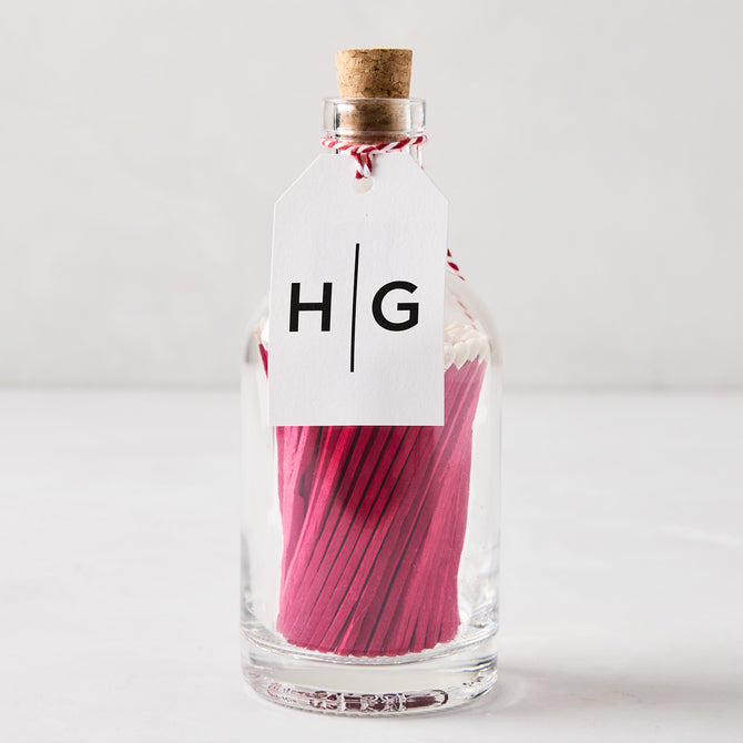 Glass bottle with cork stopper, filled with pink matches, and a white HG tag.