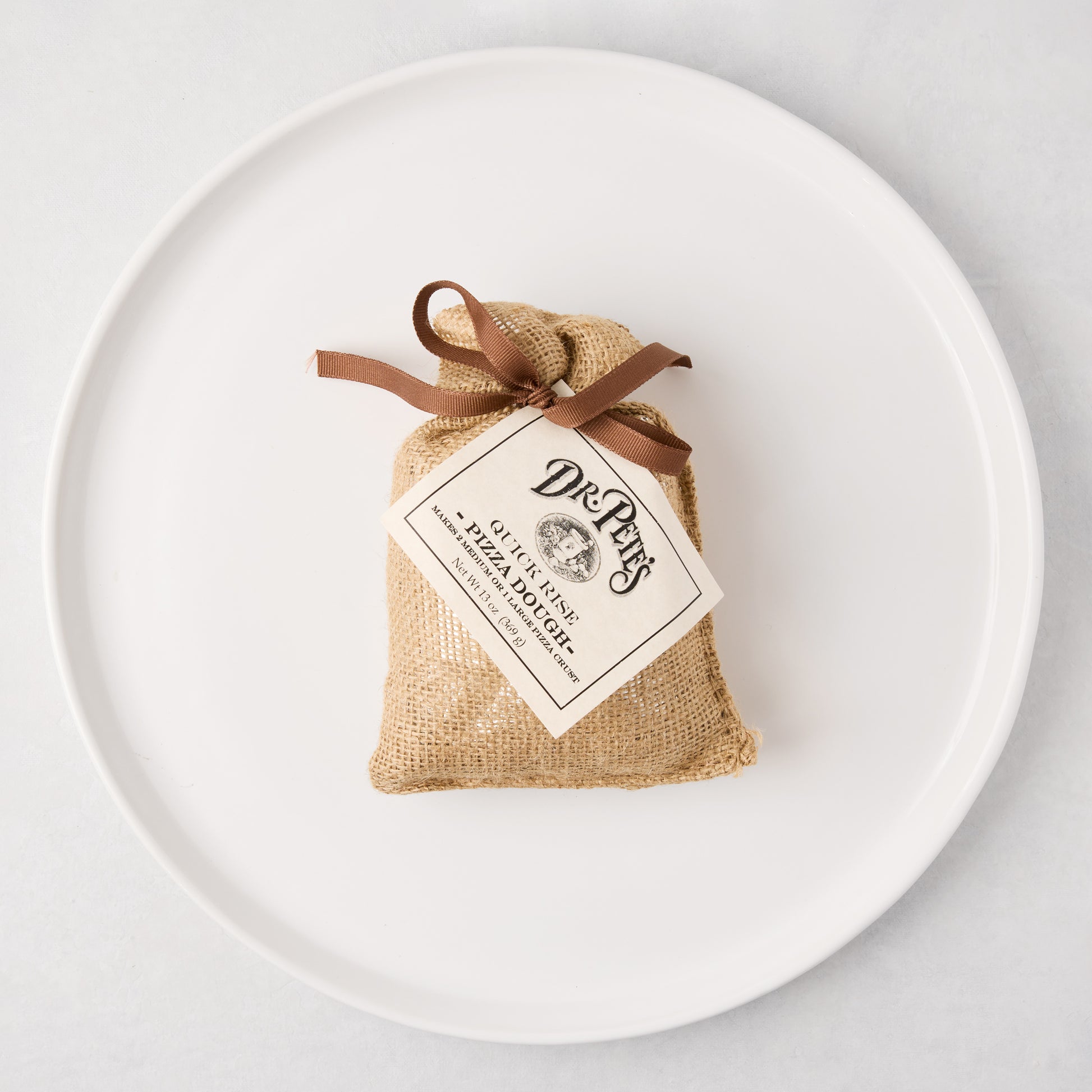 Dr. Pete's Quick Rise Pizza Dough in a small burlap bag with a brown ribbon, on a white ceramic plate.