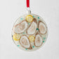 Round glass ornament with five oysters on ice, lemon slices, and blue accents, hanging from a red ribbon.