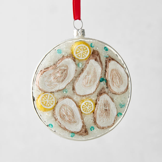 Round glass ornament with five oysters on ice, lemon slices, and blue accents, hanging from a red ribbon.