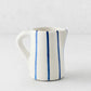 Small Portofino ceramic pitcher with white glaze, hand-painted blue vertical stripes, curved handle, and spout.