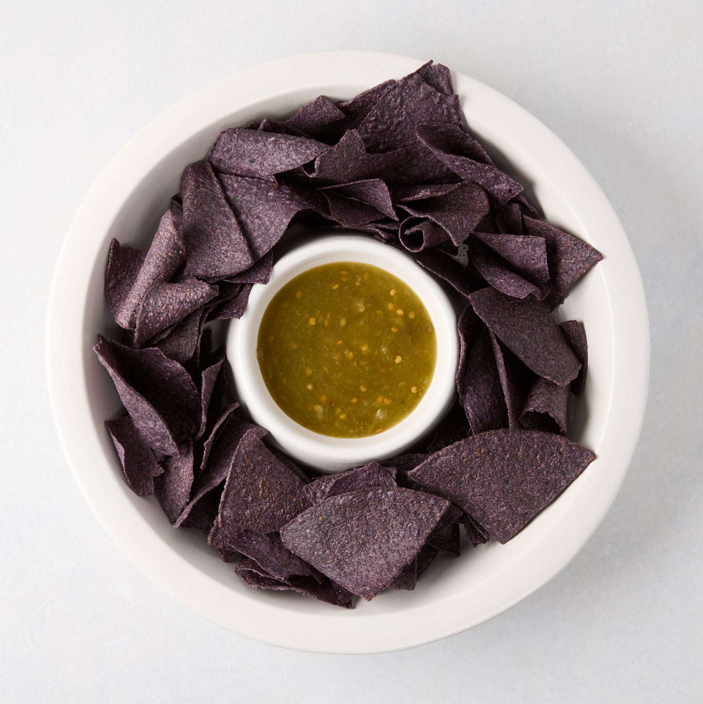 White Ravello stoneware chip and dip platter with built-in bowl, filled with blue chips and green salsa.