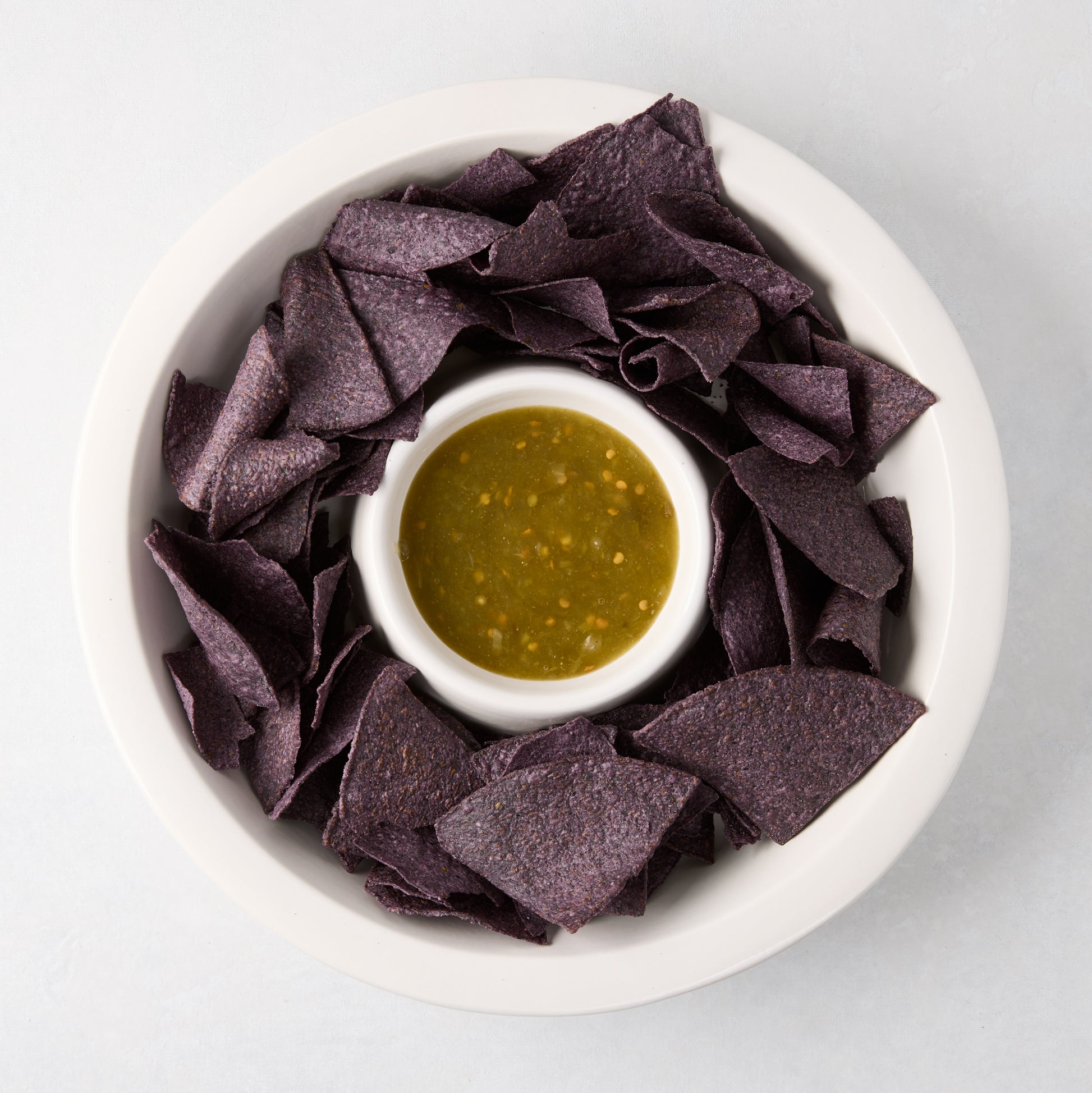 White Ravello stoneware chip and dip platter with built-in bowl, filled with blue chips and green salsa.