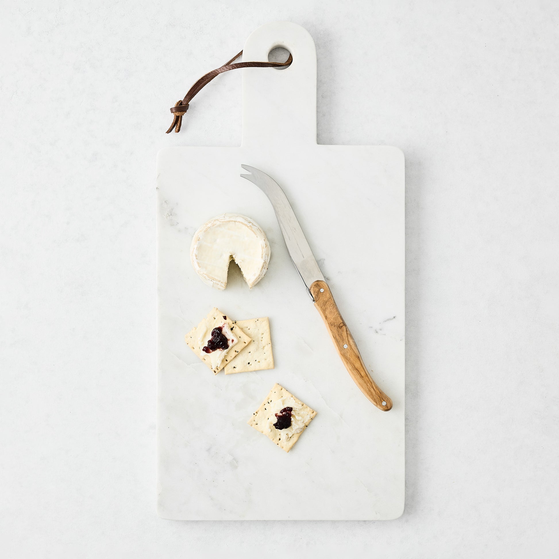 White rectangular marble serve board with handle, leather strap, cheese, crackers, and knife.