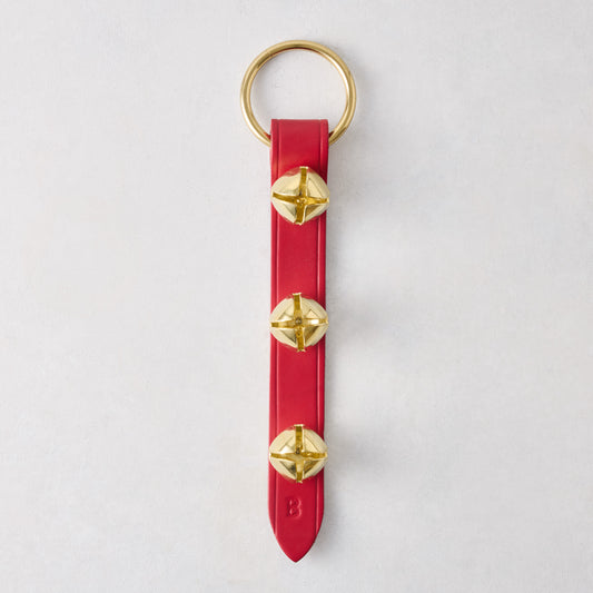 Three gold-toned jingle bells attached to a vertical red leather strap with a brass ring on a white background.