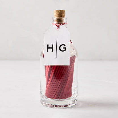 Glass bottle with cork stopper filled with red matches with white tips, tied with a white "HG" tag.
