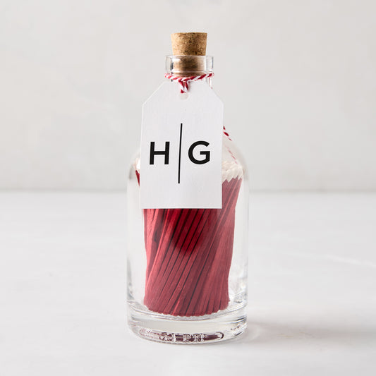 Glass bottle with cork stopper filled with red matches with white tips, tied with a white "HG" tag.