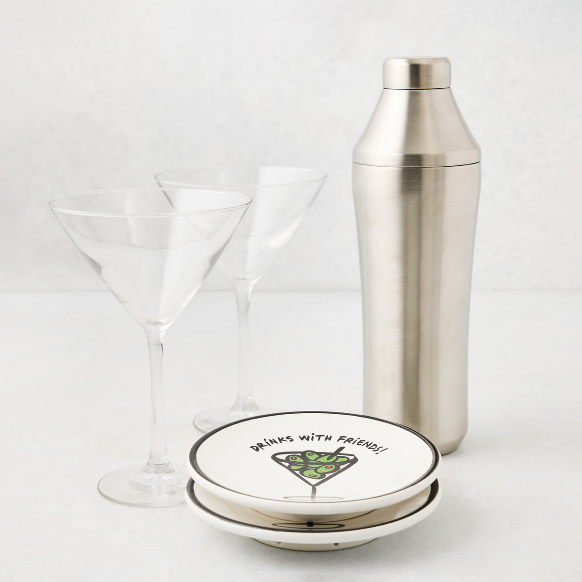 Two clear martini glasses, a brushed stainless steel cocktail shaker, and two small white plates with olive illustrations.