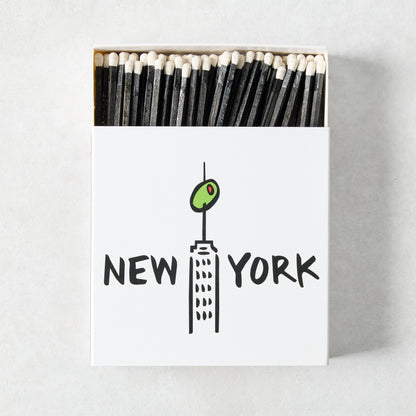 Oversized matchbox with black matches and a white box featuring a New York skyscraper illustration with an olive.
