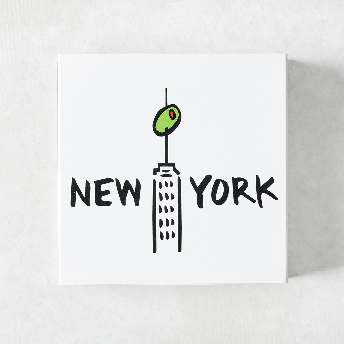 White square matchbox featuring a black line illustration of a New York skyscraper topped with a green olive.