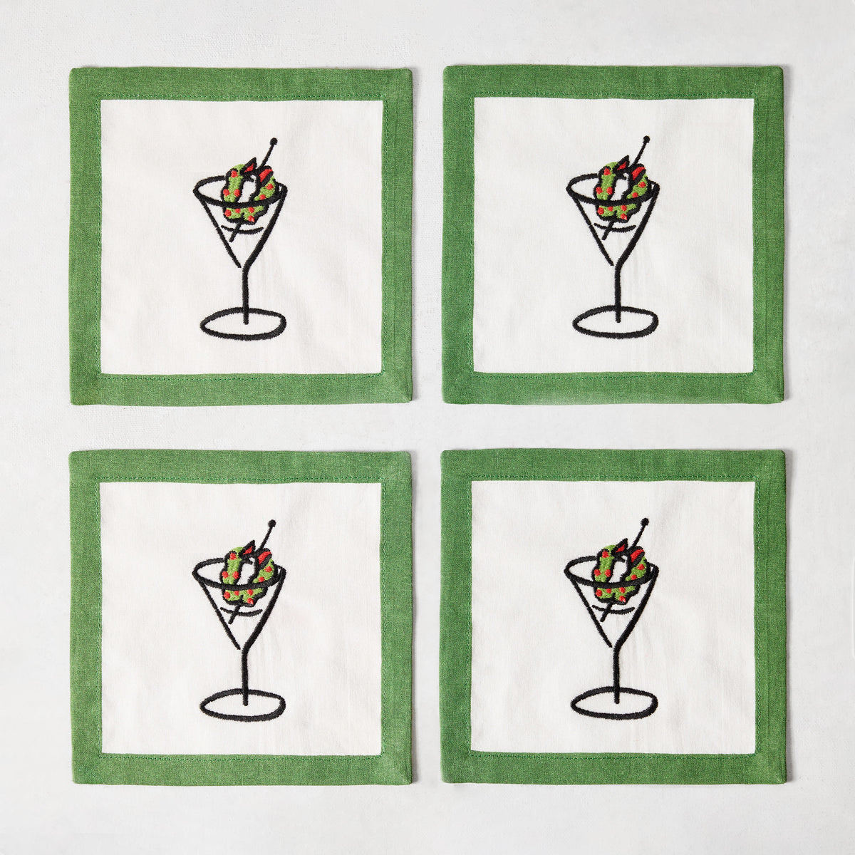 Set of four white linen coasters with green borders and embroidered martini glass illustrations with wreath garnishes.