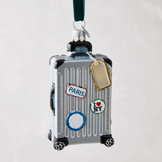 Silver glass rolling suitcase ornament with "Paris" and "I ❤️ NY" stickers, glitter details, and a gold luggage tag.