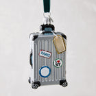 Silver glass rolling suitcase ornament with "Paris" and "I ❤️ NY" stickers, glitter details, and a gold luggage tag.
