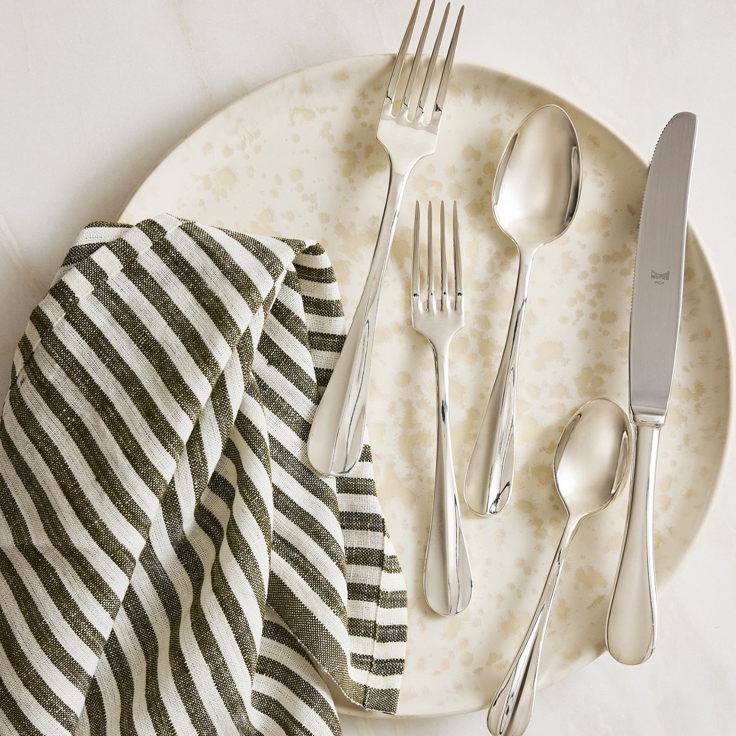 Roma Hotel 5-piece silverplate flatware set with a modern Neo-Baroque design on a speckled plate with a striped napkin.
