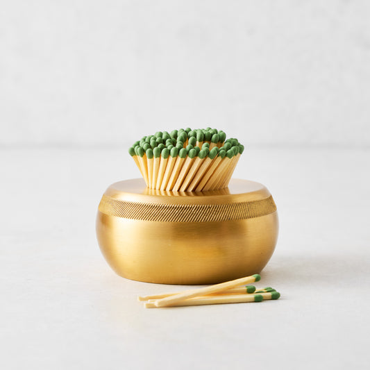 Rounded brass match striker with a textured rim, filled with wooden matches featuring green tips on a white surface.