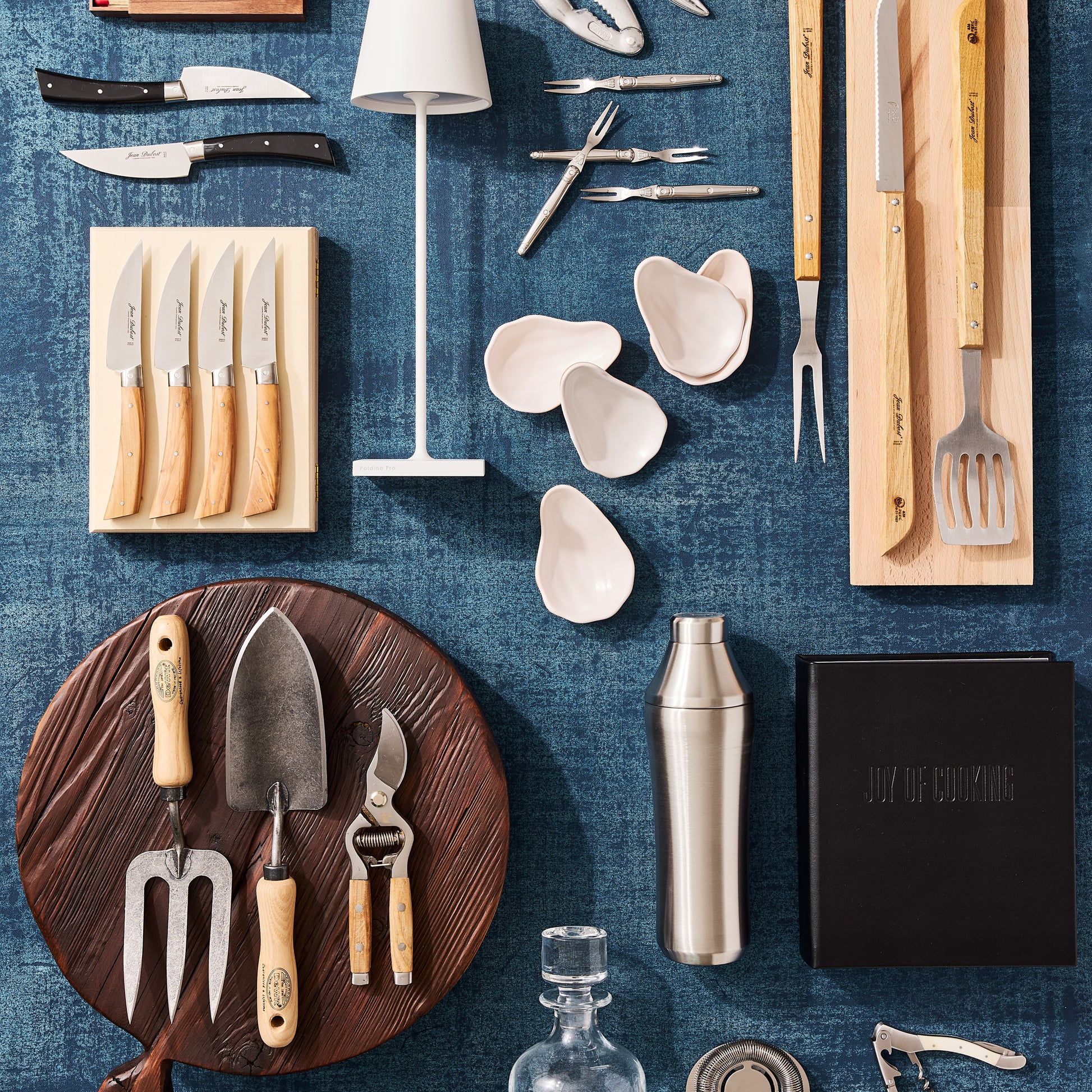 Dark brown, round, footed wood serving board with handle, garden tools, and kitchen utensils on a blue background.