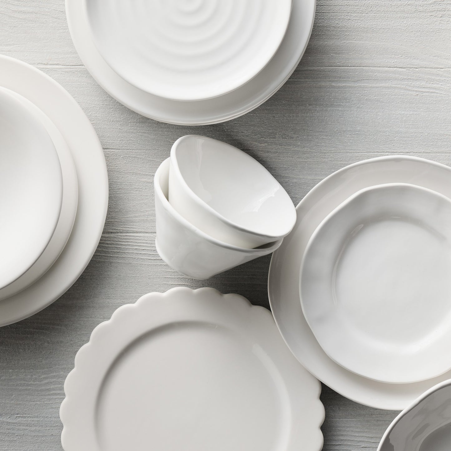 White ceramic scalloped dinner plate, salad plate, and bowls on a light wood surface.