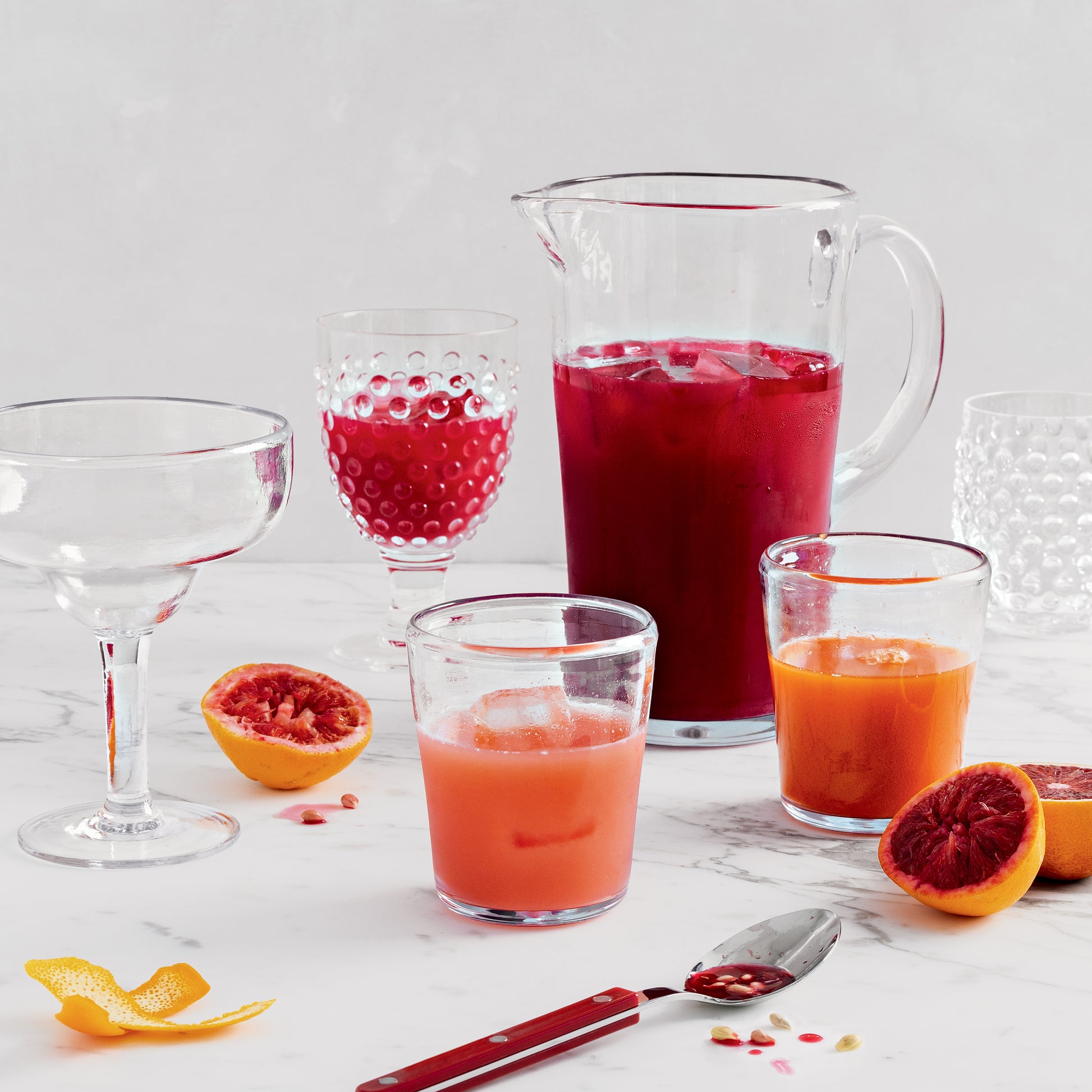 Clear acrylic Seadrift pitcher filled with red liquid, surrounded by glasses, blood oranges, and a spoon.