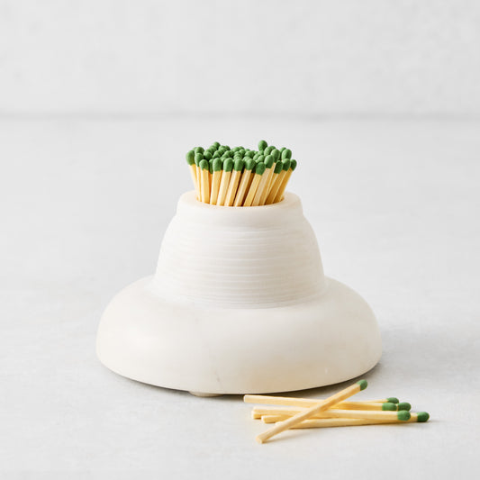 Rounded white marble match striker filled with wooden matches featuring green tips on a light surface.