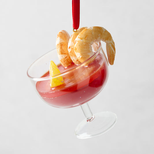 Glass shrimp cocktail ornament with red sauce, lemon wedge, and three shrimp, hanging from a red ribbon.