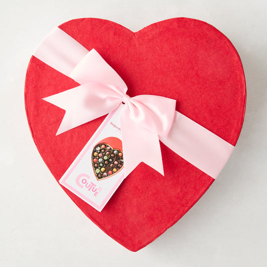 Red heart-shaped chocolate box with pink ribbon bow and "Couture" tag showing colorful chocolates.