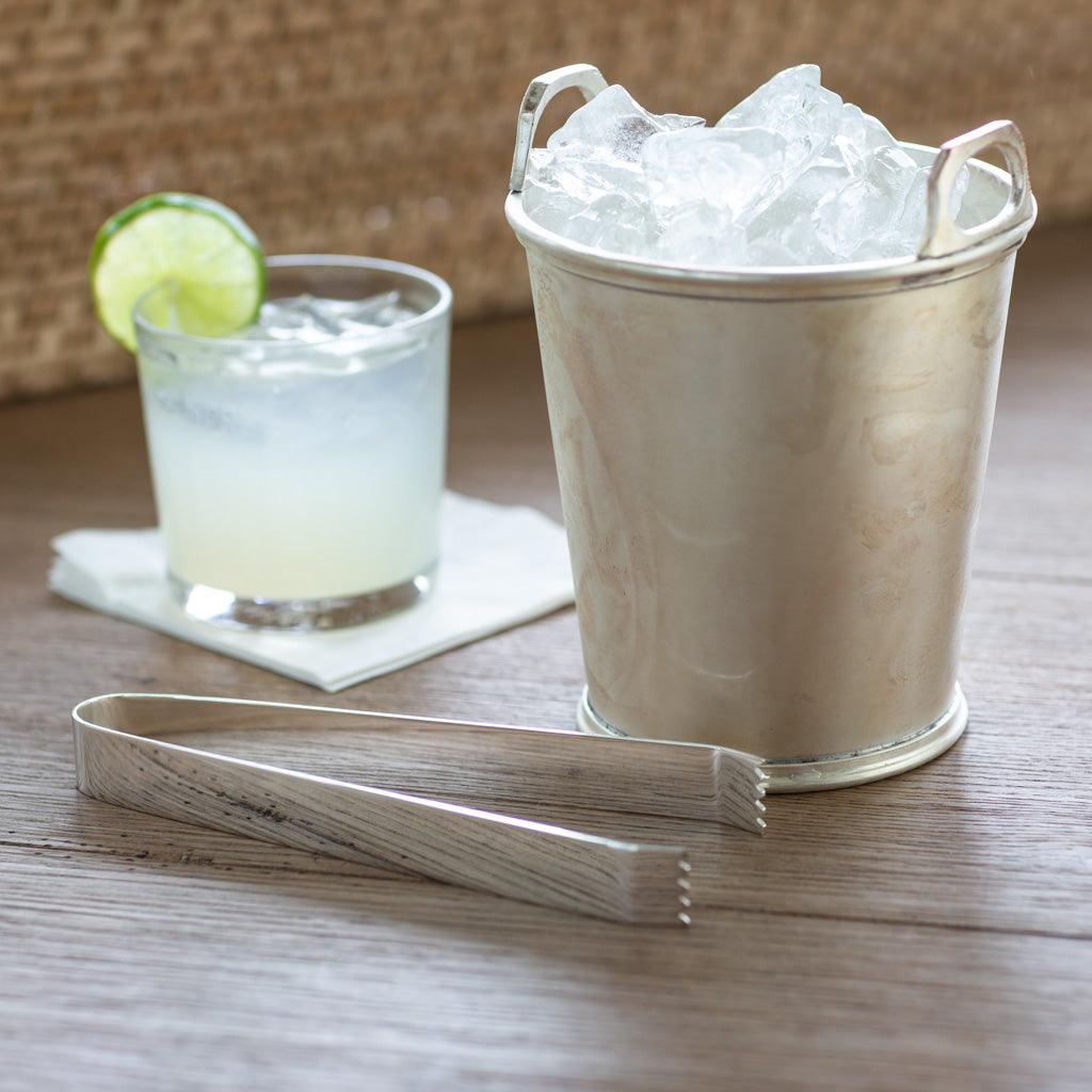 HG Silverplate Ice Tongs, silver, with a textured handle, next to an ice bucket and cocktail.