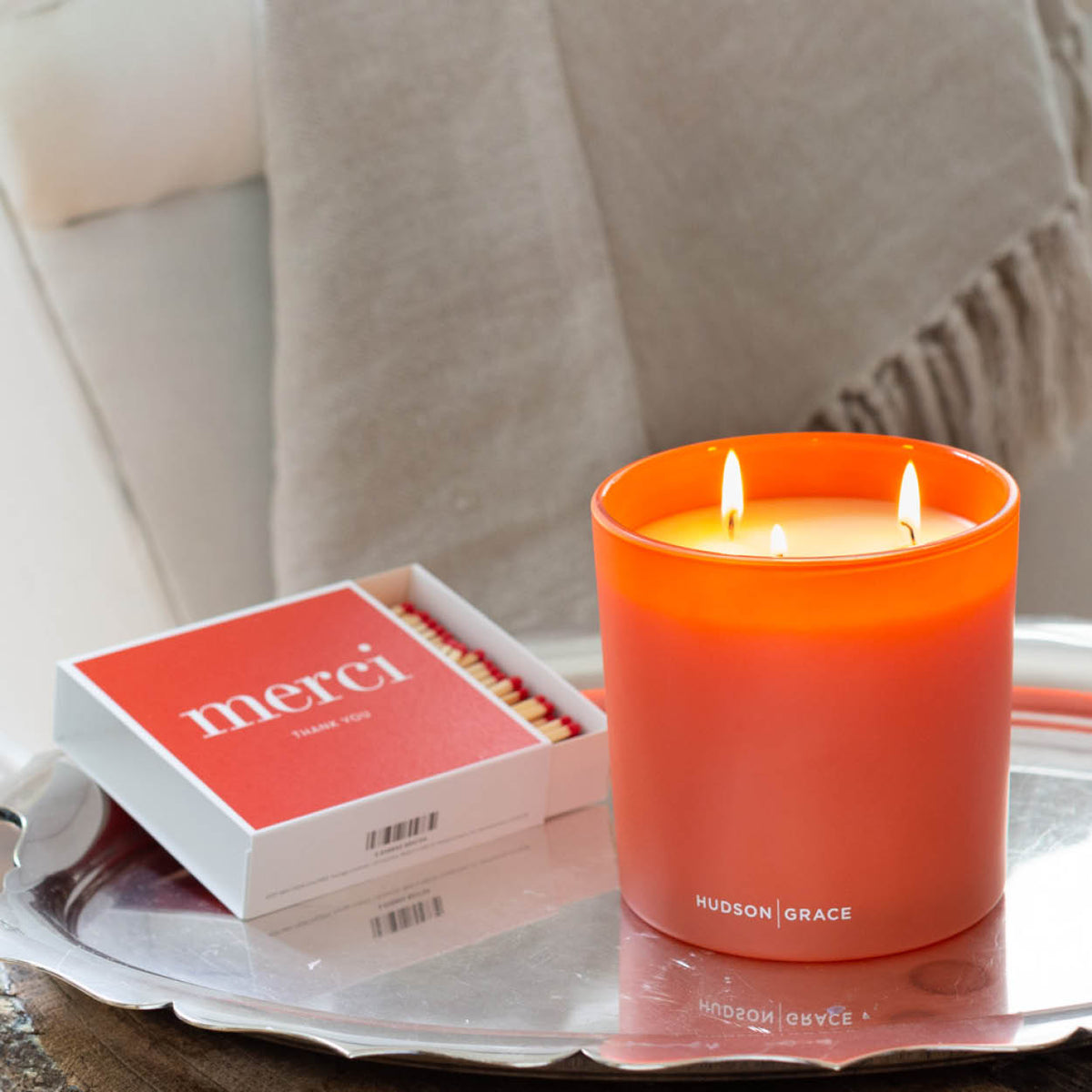 Three-wick orange candle by Hudson Grace and a box of Merci matches on a silver tray.