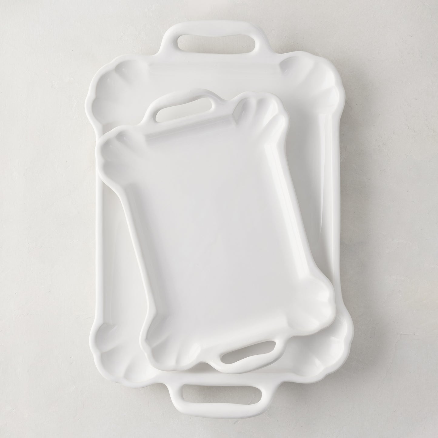 Two white ceramic Alessia serving platters, one small on top of a large, with handles and scalloped edges.