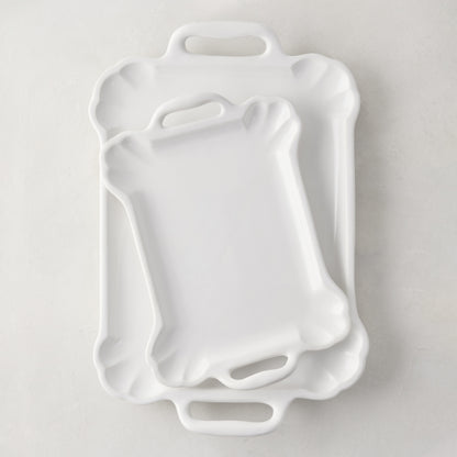 Two white ceramic Alessia serving platters, one small on top of a large, with handles and scalloped edges.