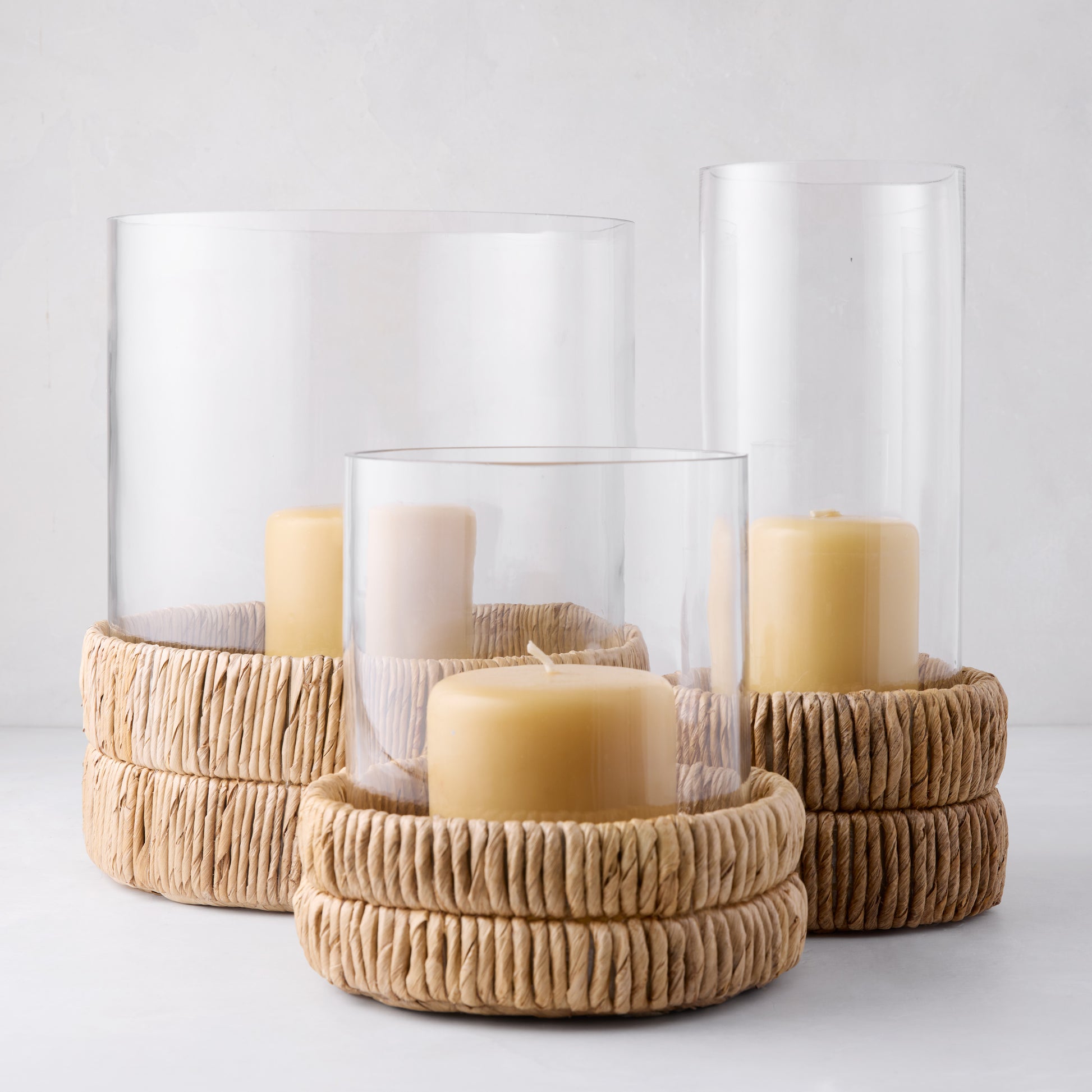 Three clear glass hurricanes with woven banana bark bases and amber candles.
