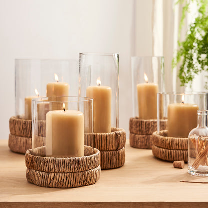 Small glass hurricane with a woven banana bark base and lit amber candle.