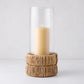 Small glass hurricane with a woven banana bark base and lit amber candle.