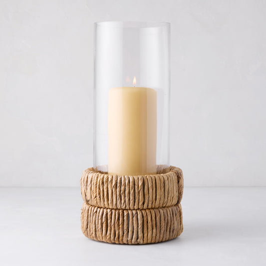 Small glass hurricane with a woven banana bark base and lit amber candle.