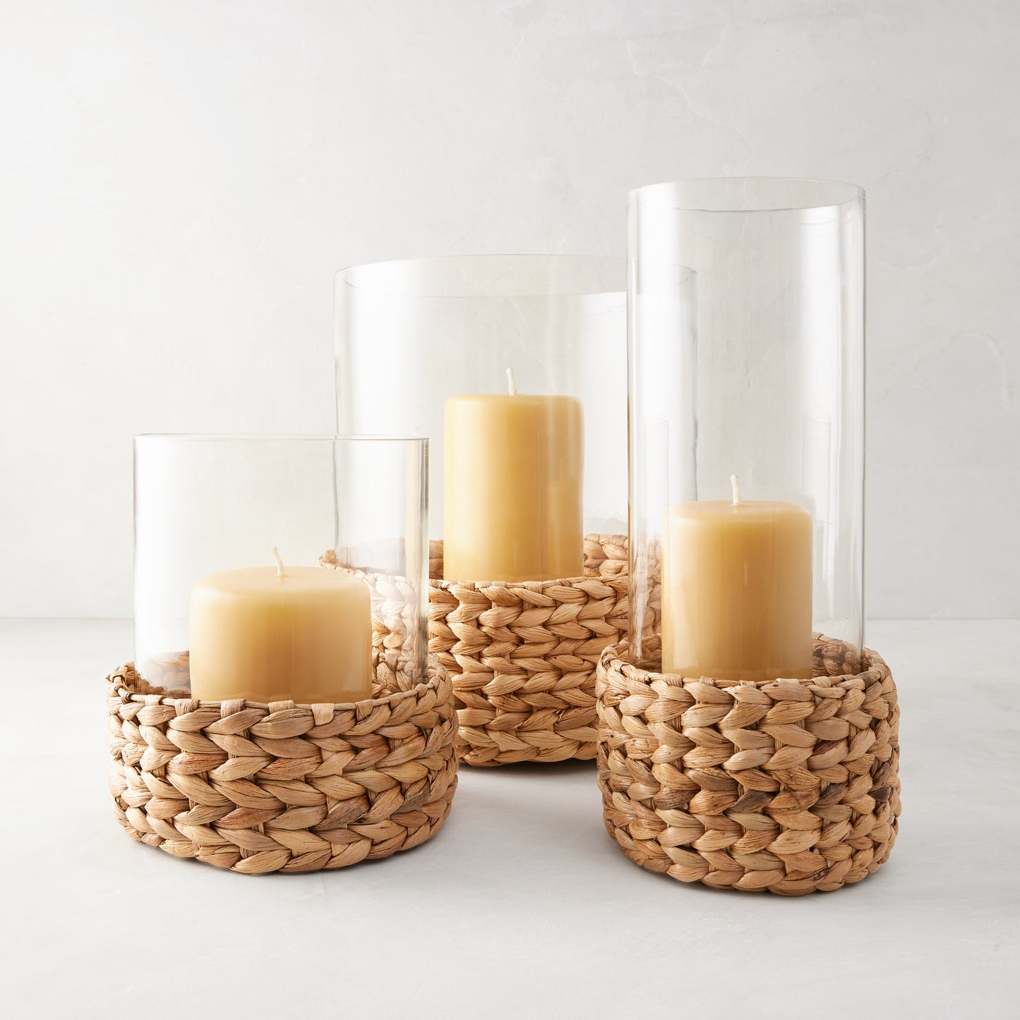 Three glass hurricanes with woven hyacinth bases and amber candles, varying in height.