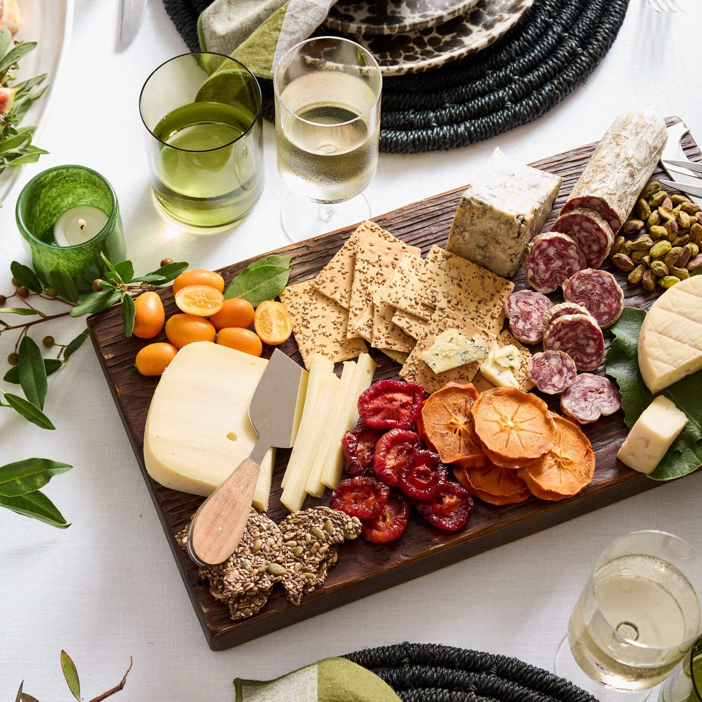 Dark wood serving plank with charcuterie, cheeses, crackers, dried fruit, and kumquats.