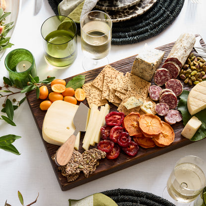 Dark wood serving plank with charcuterie, cheeses, crackers, dried fruit, and kumquats.