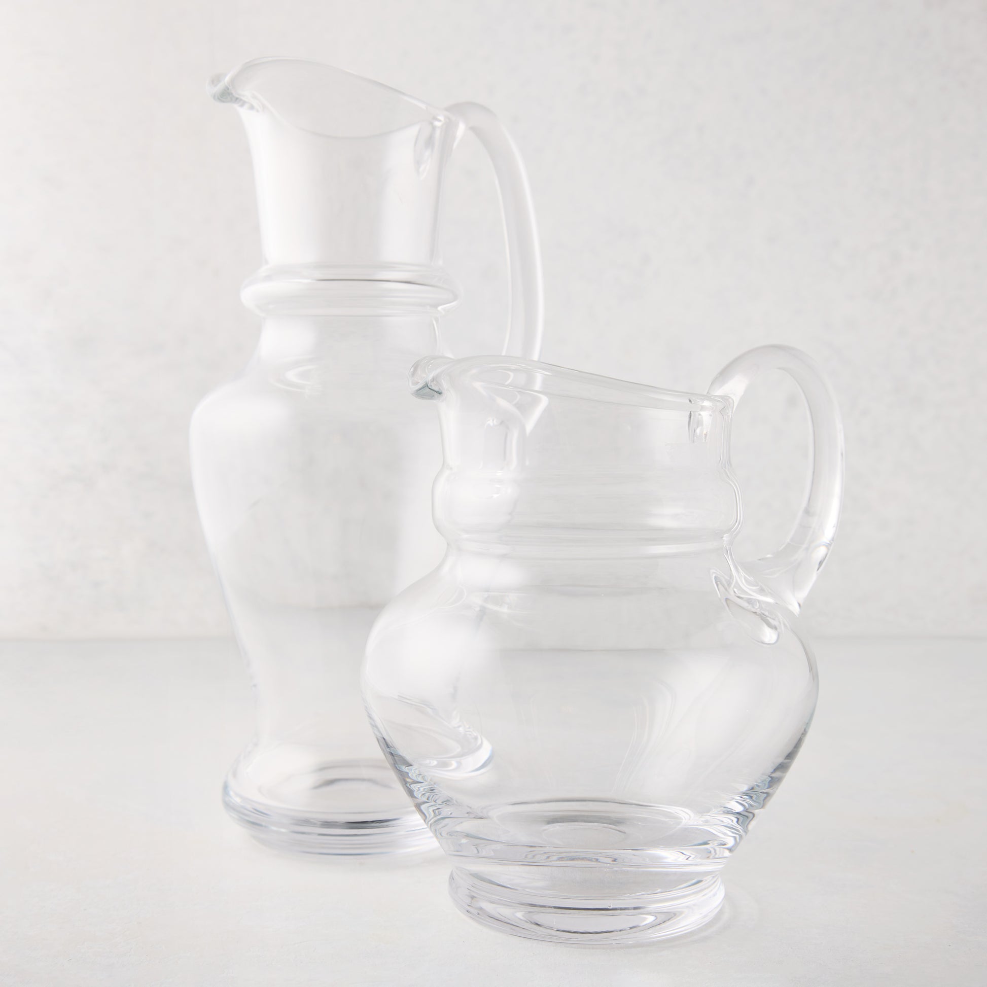 Two clear hand-blown Sutton glass pitchers, one tall and one short, with unique shapes and handles.