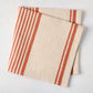 Folded Spice Orange Boat Stripe Natural Napkin, 50% cotton, 50% linen, with orange stripes on a beige base.