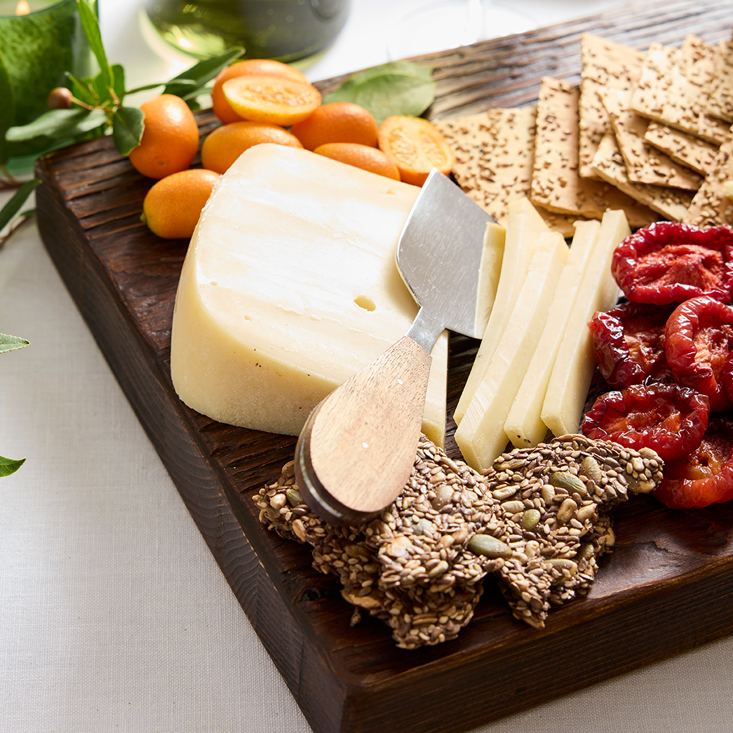 Stainless steel cheese knife with wood handle on a dark wood board with cheese, crackers, and fruit.
