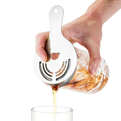 Hand holding a silver stainless steel Hawthorne cocktail strainer, pouring a drink from a glass mixer.