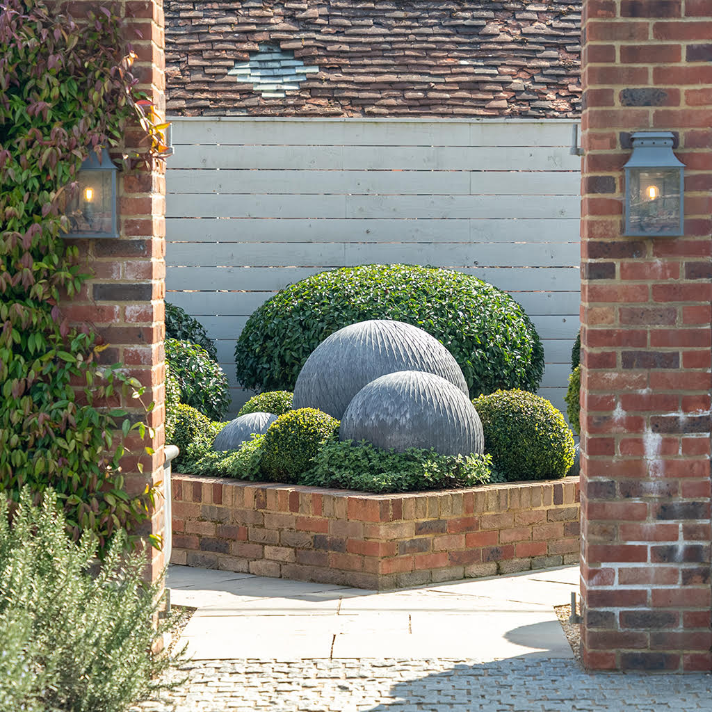 Three dark gray steel metal garden leaf balls of varying sizes in a brick planter with green plants.