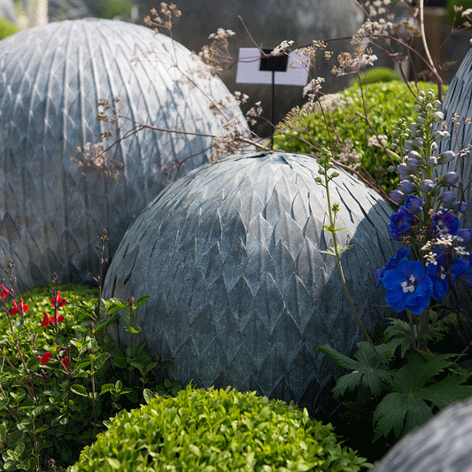 Three large, round steel garden leaf balls with layered, hand-welded metal leaves, nestled in green plants.