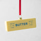 Yellow glass stick of butter Christmas tree ornament with blue "BUTTER" text and red ribbon.