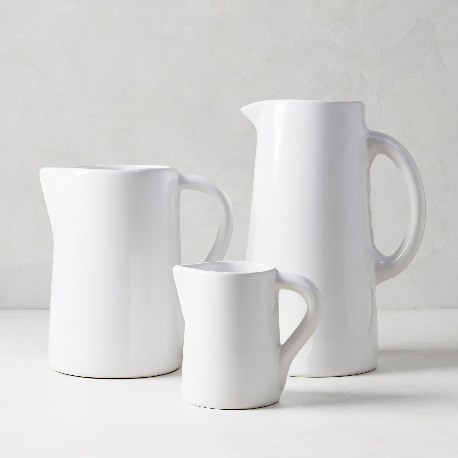 Three handmade white ceramic pitchers in varying sizes, with large handles and spouts.