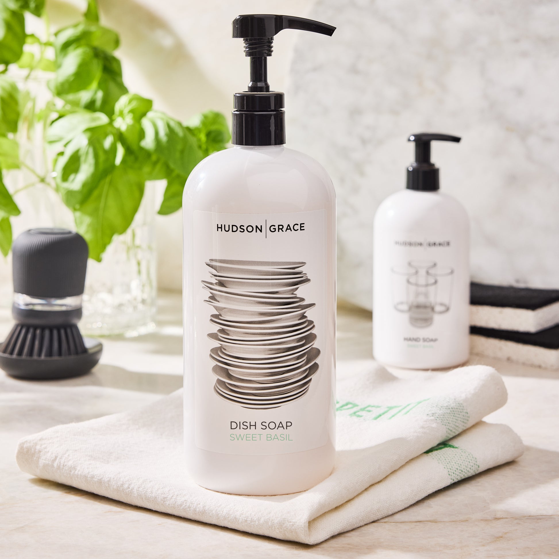 White HG Sweet Basil Dish Soap bottle with black pump, stacked dishes graphic, on a white towel.