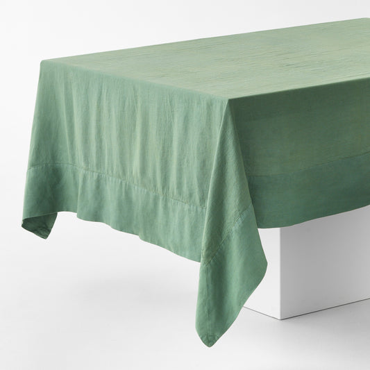 Sage green hand-dyed linen tablecloth with a wide border and mitered corner detailing draped over a white table.