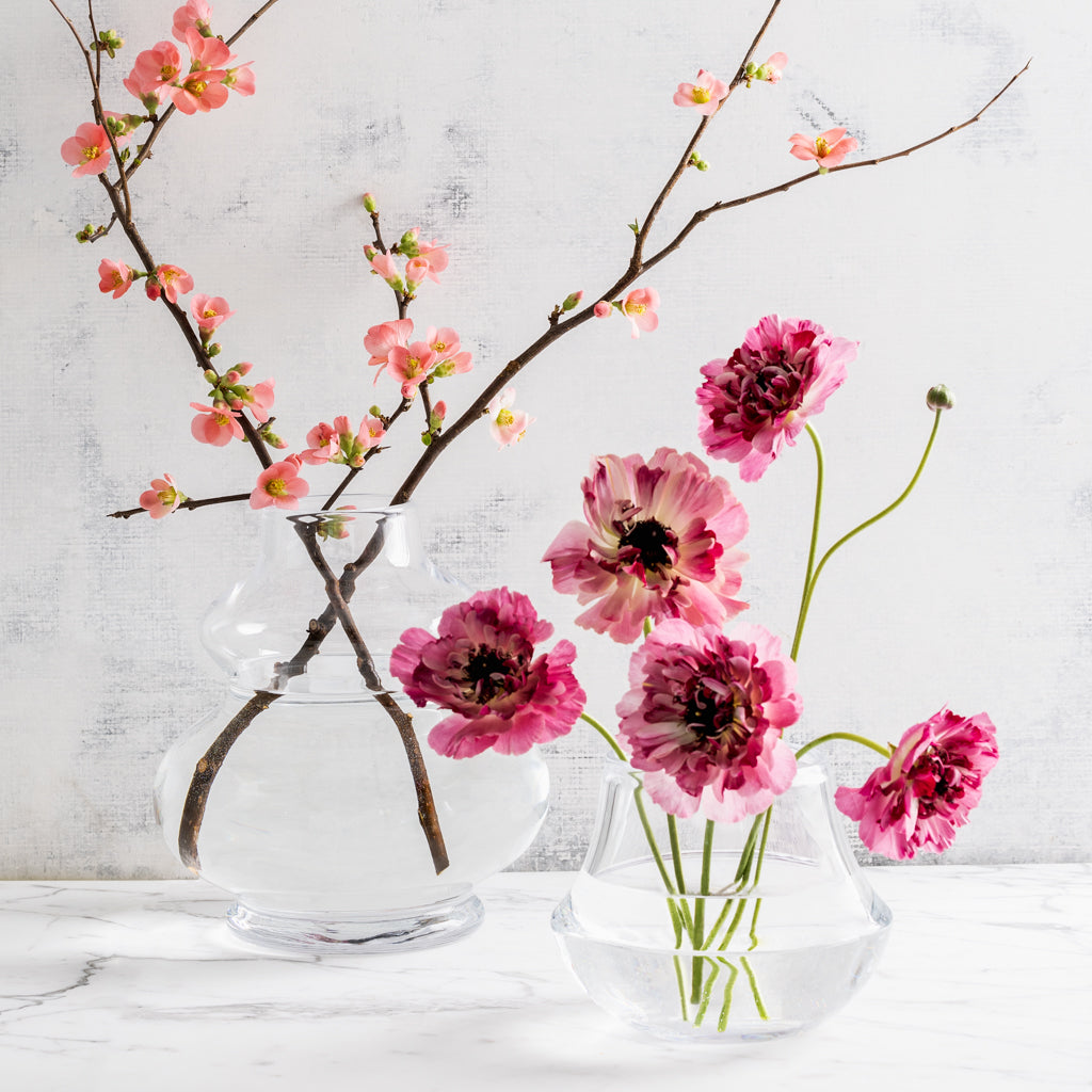 Two clear Sylvia glass vases, one tall with pink flowering branches, one short with pink ranunculus.
